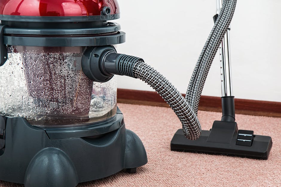 A close-up view of a vacuum cleaner with a yellow canister and a black hose, positioned on a traditional floral-patterned carpet in a living room setting. The room features a wooden floor with a mediu